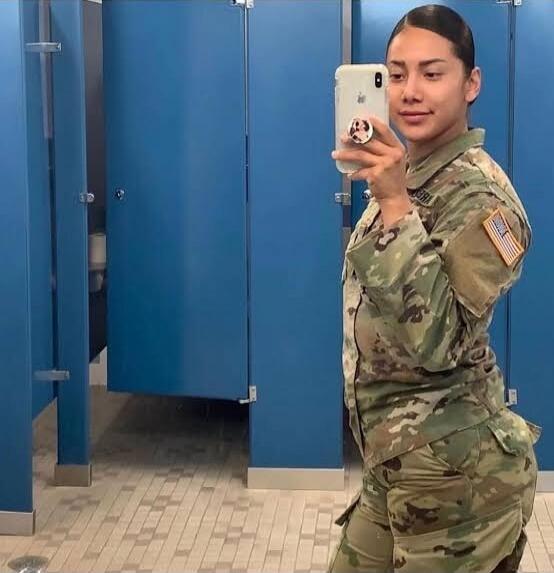 A female soldier in camouflage uniform taking a mirror selfie in a restroom/locker room.