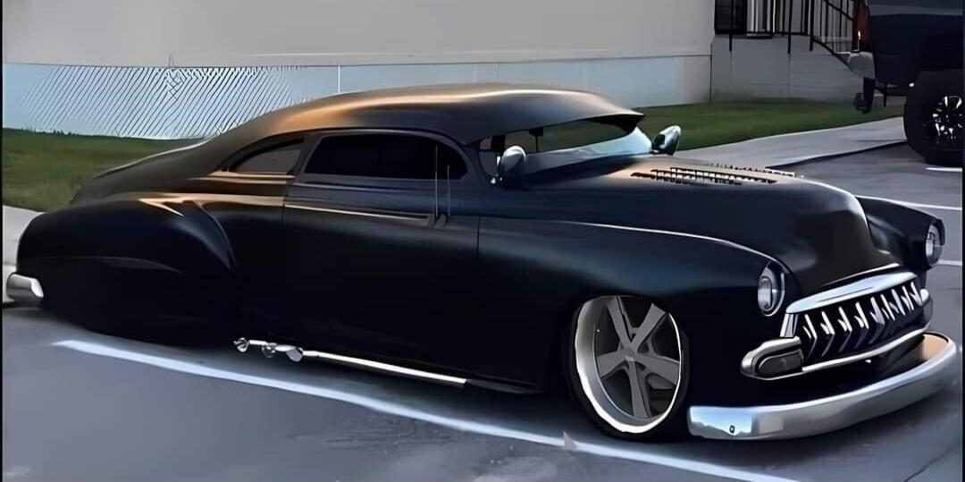 A custom black classic car, possibly a Chevrolet Fleetline or similar model from the late 1940s or early 1950s, with a chopped top, lowered suspension, and custom wheels, parked on asphalt.