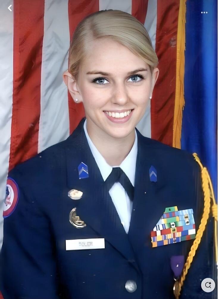 A young woman in a dark blue military or cadet uniform, white shirt, and black tie, smiling at the camera. She has blonde hair and blue eyes. Various pins, ribbons, and a name tag reading 'TOLER' are visible on her uniform. An American flag and another blue flag are in the background.