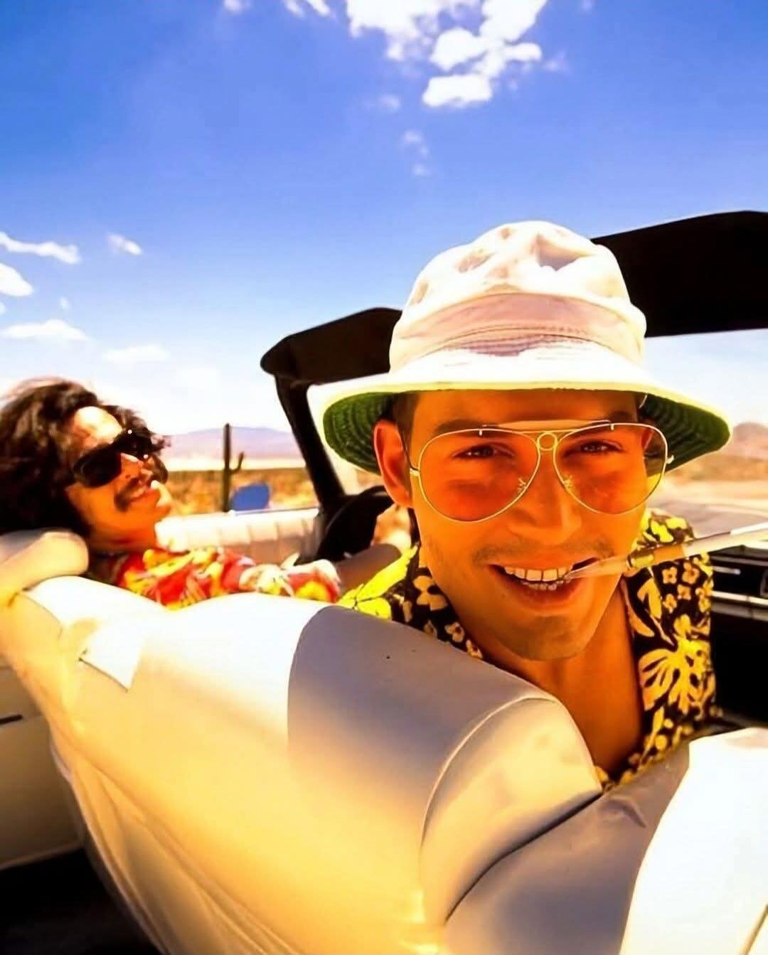 Two men in a convertible car, driving through a desert landscape under a bright blue sky. The man in the foreground wears a white bucket hat and yellow-tinted aviator sunglasses, smiling with a cigarette in his mouth. The other man is visible in the background, also wearing sunglasses and smiling.