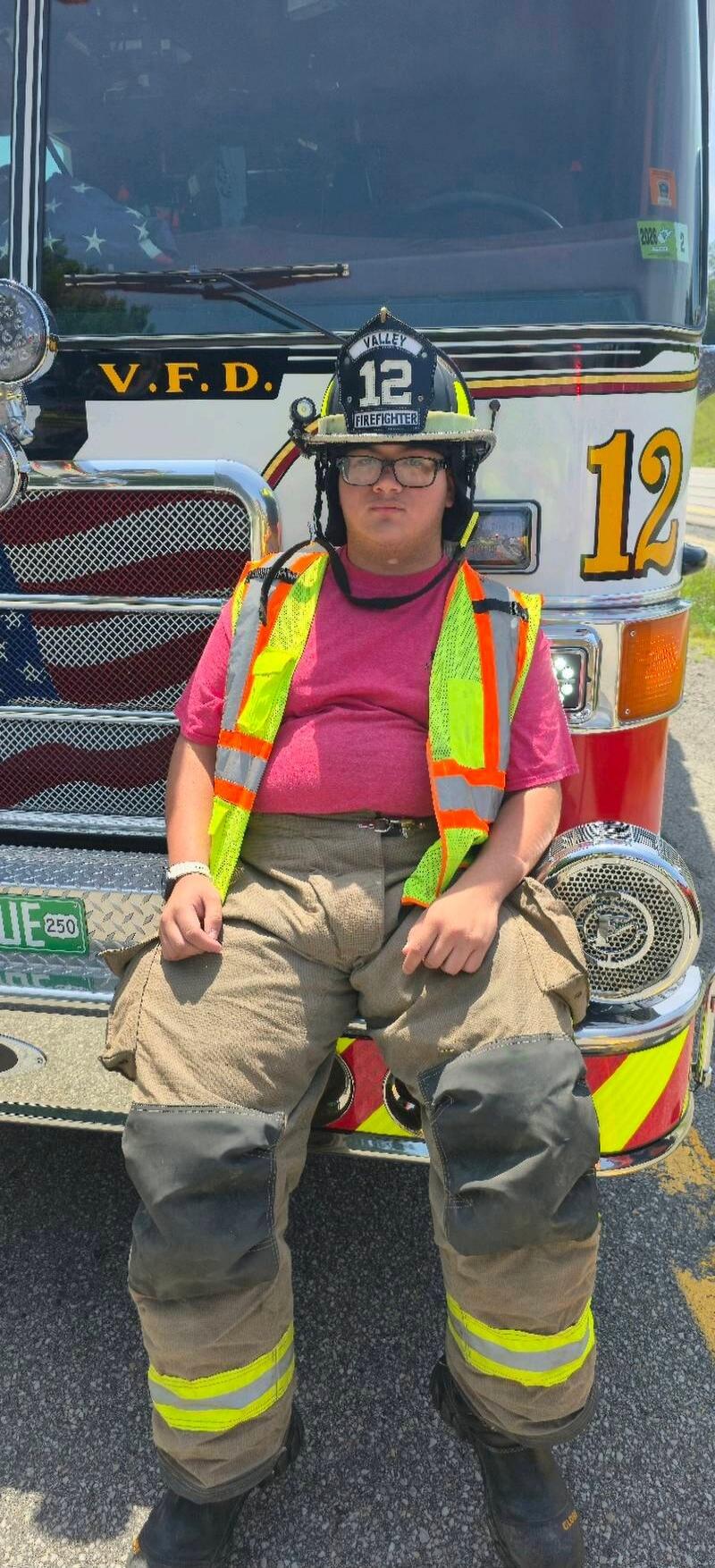 A person wearing a firefighter helmet with 'VALLEY 12 FIREFIGHTER' written on it, a high-visibility vest, and firefighter pants, sitting on the front bumper of a fire truck. The truck has 'V.F.D.' and '12' visible, and an American flag pattern on its grille.