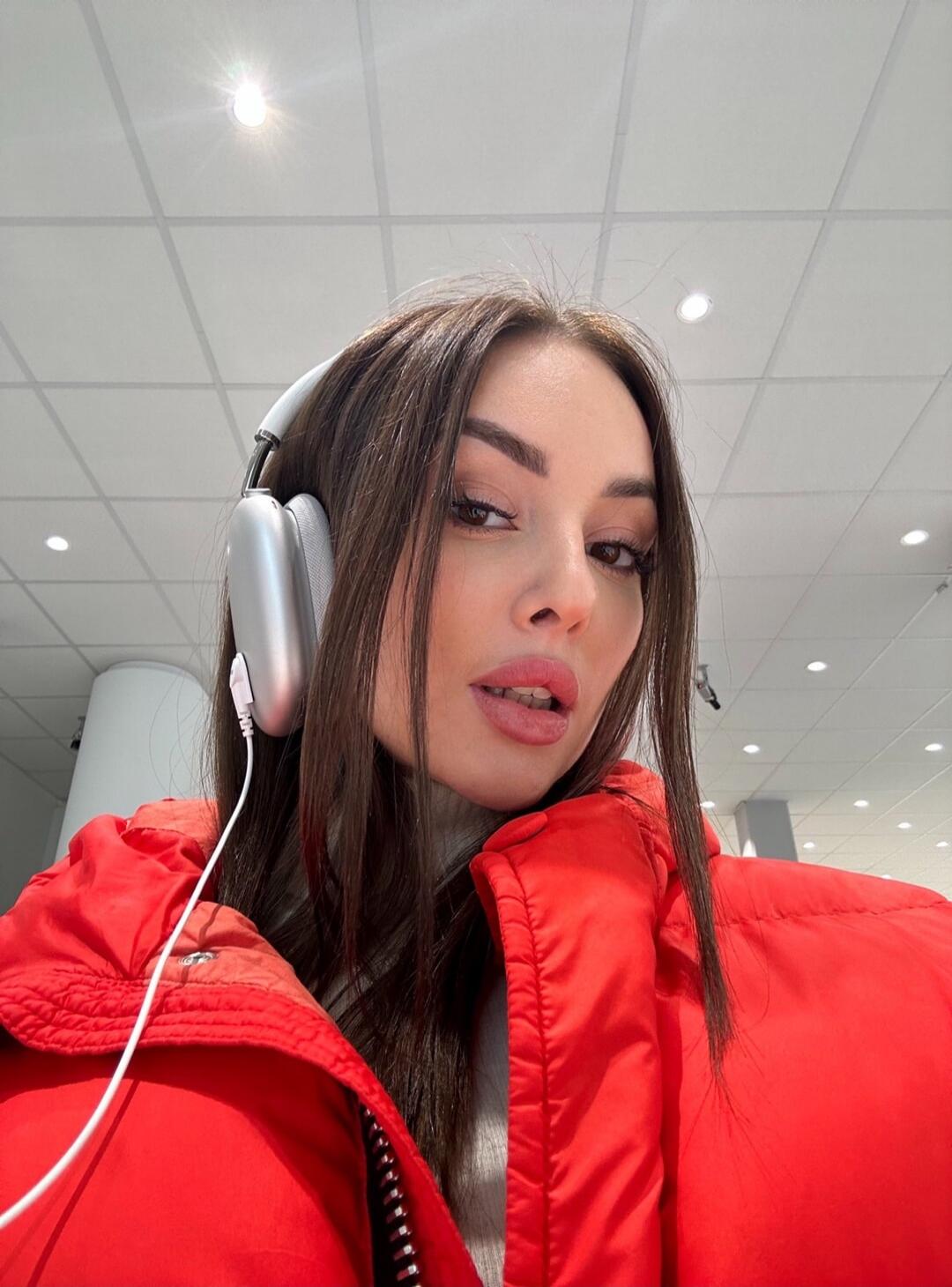 A young woman with long dark hair, wearing a red puffer jacket and silver over-ear headphones, looking directly at the camera in an indoor setting.