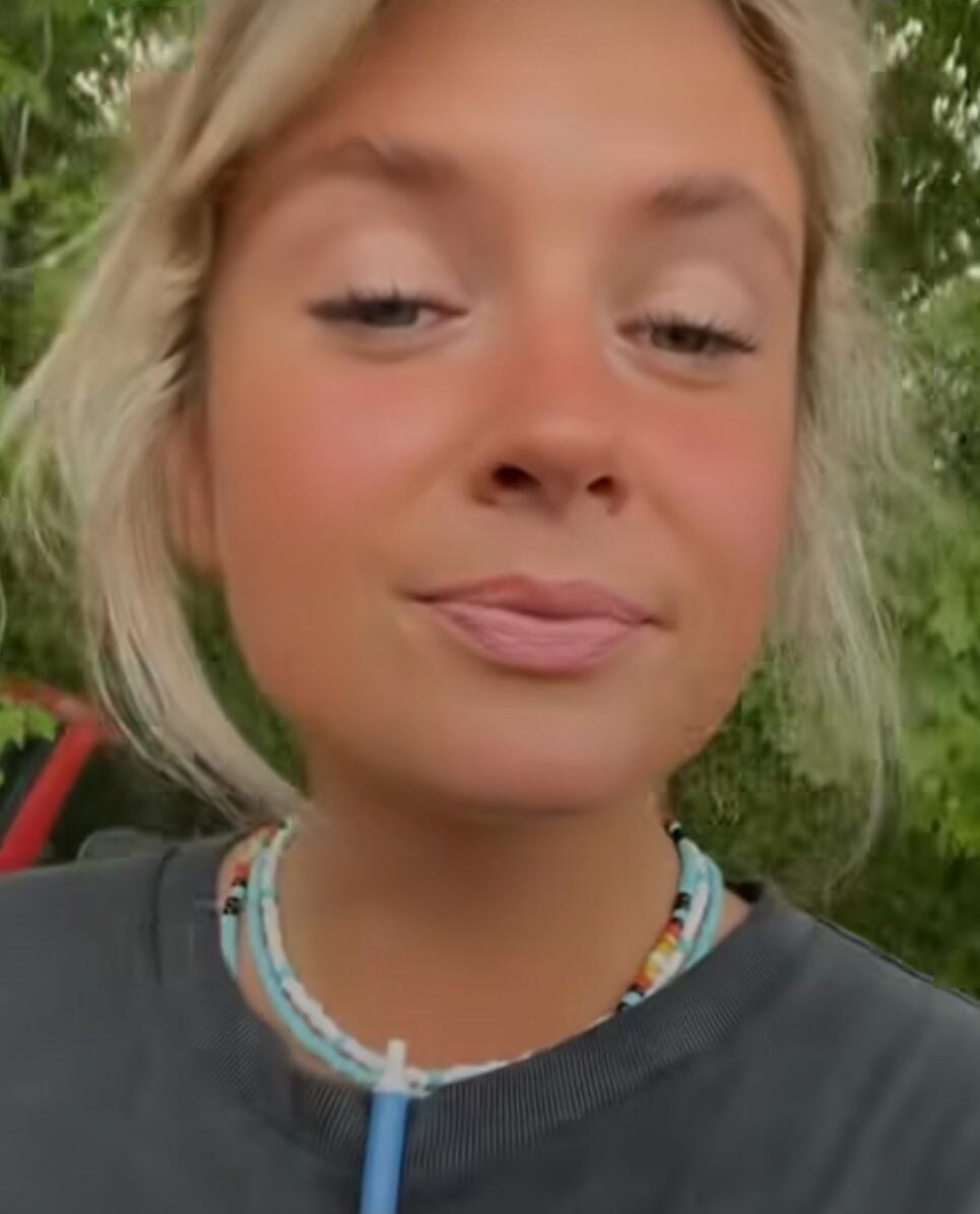A close-up portrait of a young woman with blonde hair outdoors, smiling softly.