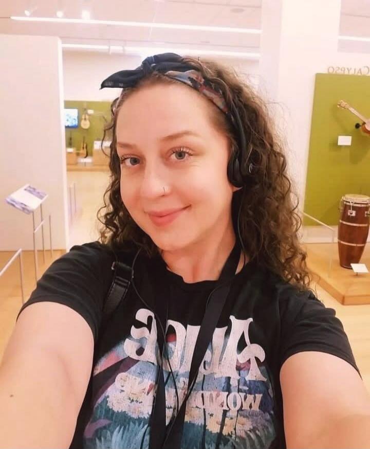 A woman with curly hair, wearing a headband and headphones, takes a selfie in what appears to be a museum or exhibition hall. Musical instruments, including a conga drum and guitars, are visible in the background.