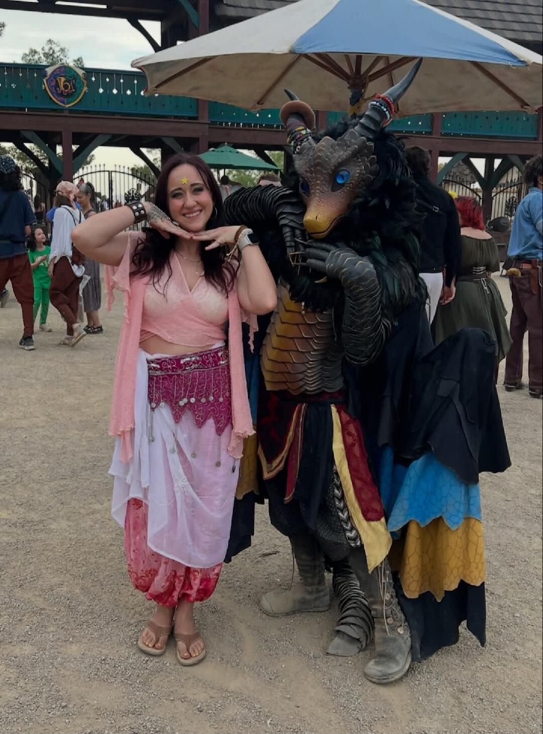 A woman in a pink and white costume with a star on her forehead poses happily next to a person in a detailed dragon costume. They are at an outdoor festival or fair, with other people and structures in the background.