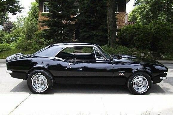 Black classic muscle car parked on a driveway. Looks like a late 1960s Chevrolet Camaro with polished wheels and chrome details.