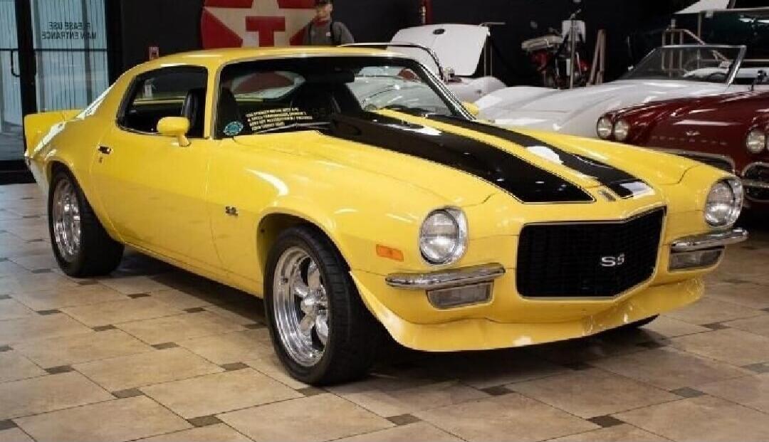 A classic yellow Chevrolet Camaro Z28 with black racing stripes, parked in a showroom or museum. Other vintage cars are visible in the background.