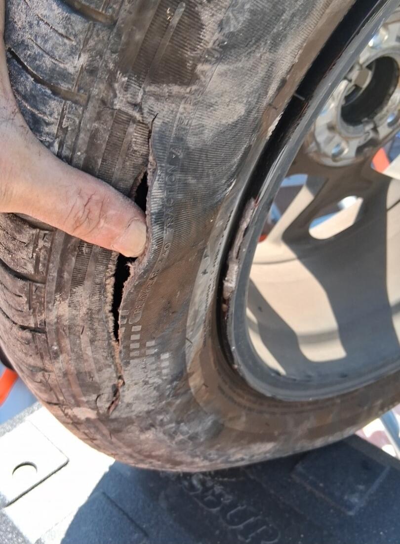 A close-up shot of a severely damaged car tire with a large tear in its sidewall, exposing the internal structure. A hand is visible, pointing at the damage. The tire appears dirty and worn, and the rim is also visible. Text on the tire includes 