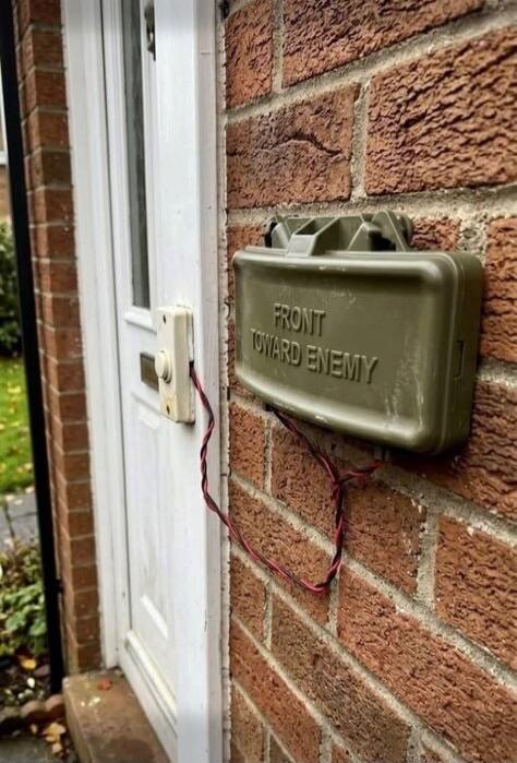 A doorbell is wired to a military-style green box on a brick wall. The box has the text 'FRONT TOWARD ENEMY' written on it, mimicking a Claymore mine. This appears to be a humorous prank or a creative doorbell setup.