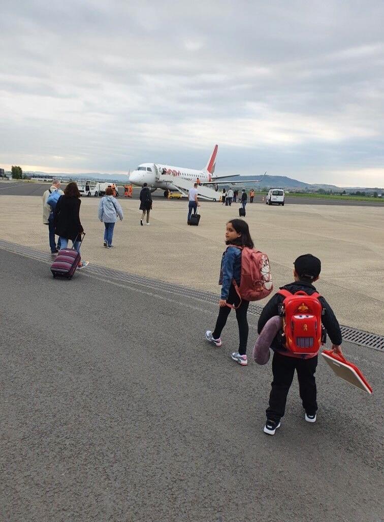 People walking on an airport tarmac towards an airplane. Two children are in the foreground, one with a pink backpack and another with a red 'Cars' backpack, holding a red tray. An airplane with 'JAT Airways' written on its side is visible in the background. The sky is cloudy.