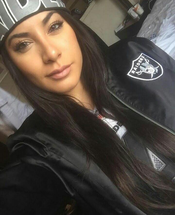A young woman wearing a black jacket with a Raiders logo and a cap, taking a selfie.