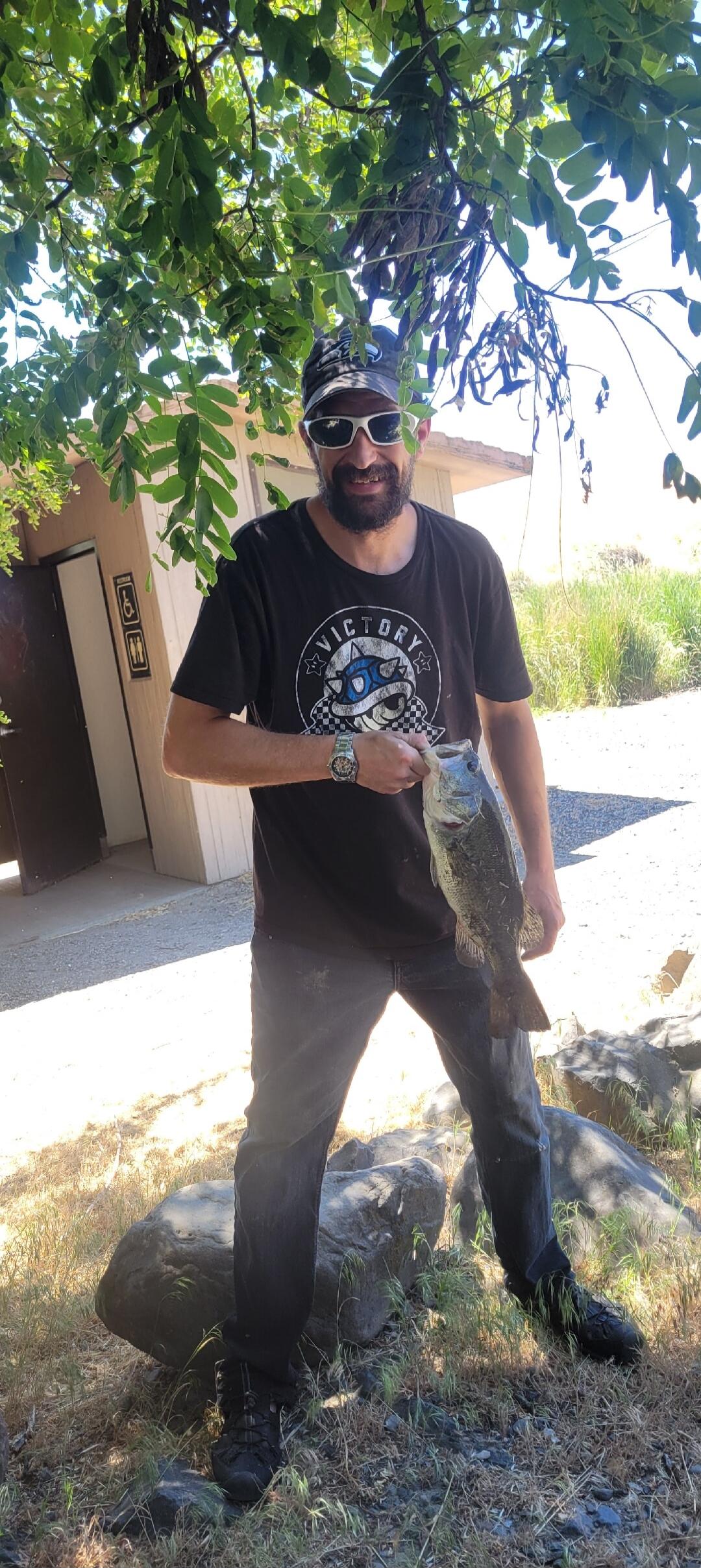 A man holding a large fish, possibly a bass, under a tree. He is wearing a t-shirt with 'VICTORY' written on it. In the background, there is a building with restroom signs.