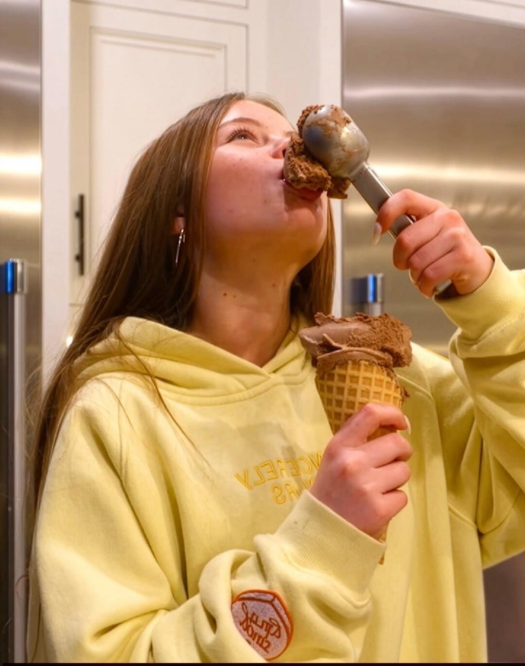 A young woman is enthusiastically eating chocolate ice cream from a cone and a scoop. She is wearing a yellow hoodie.