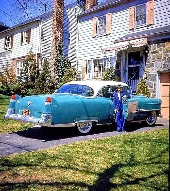 A retro turquoise car with a white roof parked in a driveway in front of a suburban house. A person in a blue suit is standing by the car.