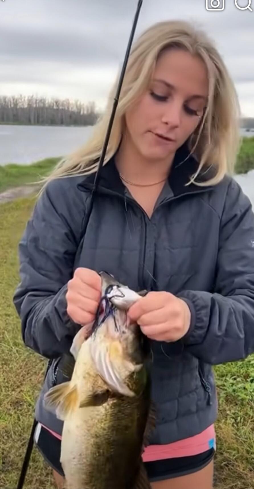 A woman wearing a gray jacket is holding a large fish by the mouth near a lakeside.