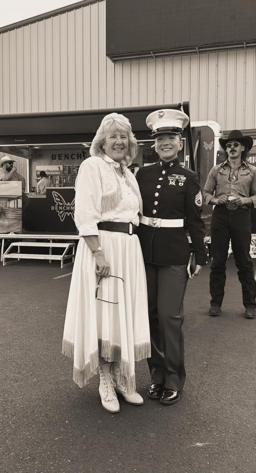 BENCHMADE. Two women, one in a white dress with fringe and cowboy boots, and another in a US Marine Corps uniform, are smiling and standing together. In the background, there is a booth with 'BENCHMADE' visible and two men, one wearing a cowboy hat.