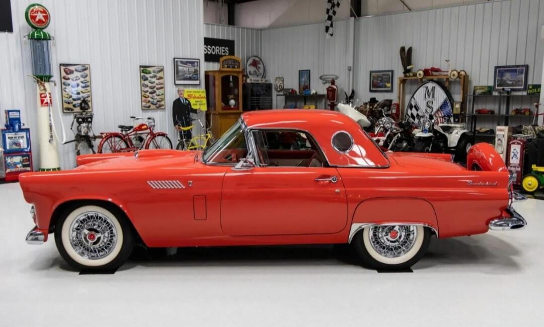 Red vintage car on display in a showroom with motorcycles and posters in the background.