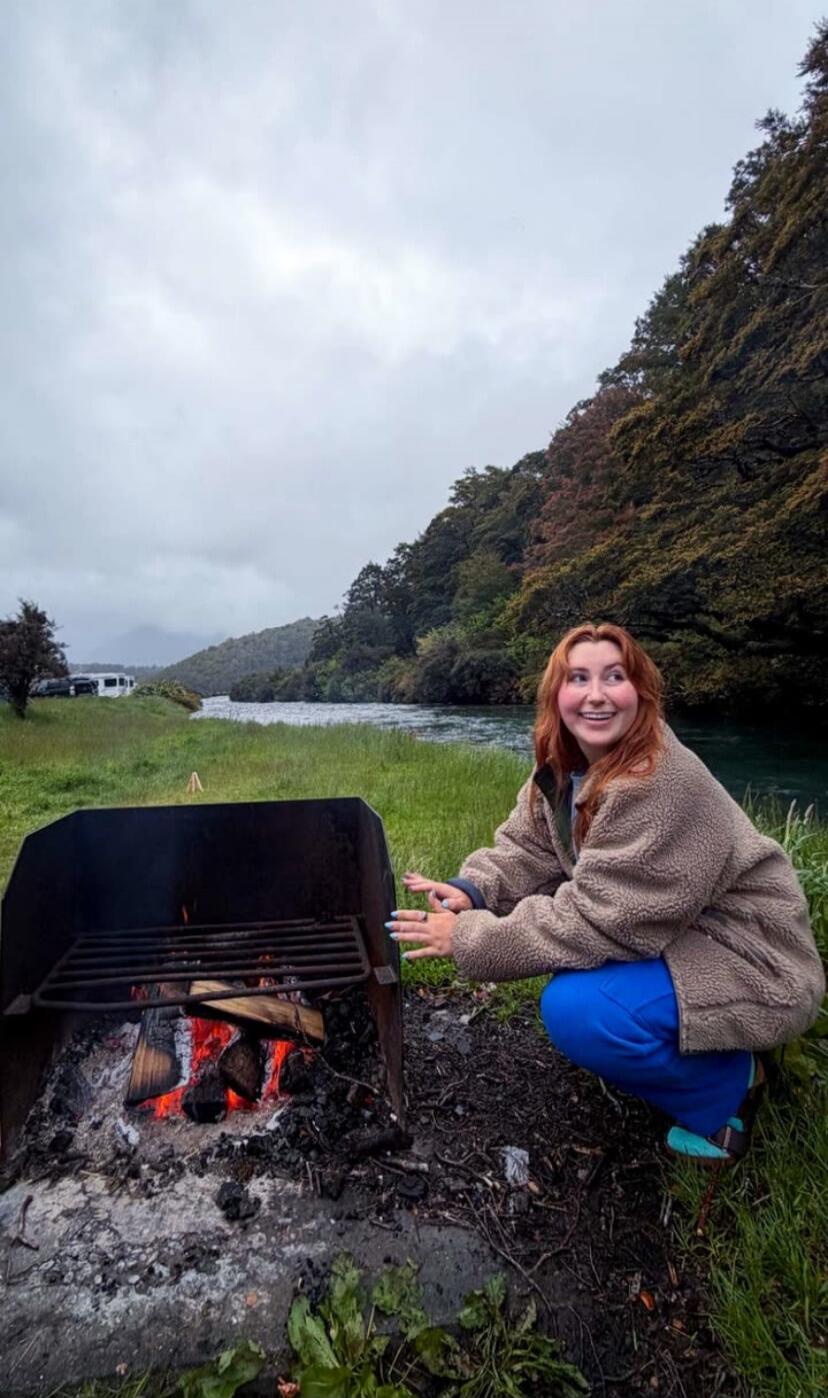 A young woman is squatting next to a campfire, smiling and looking to the side. She is wearing a fluffy brown jacket and blue pants. The campfire is burning with logs and embers. In the background, there is a river and a forested hill under a cloudy sky. There are also some RVs parked in the distance.