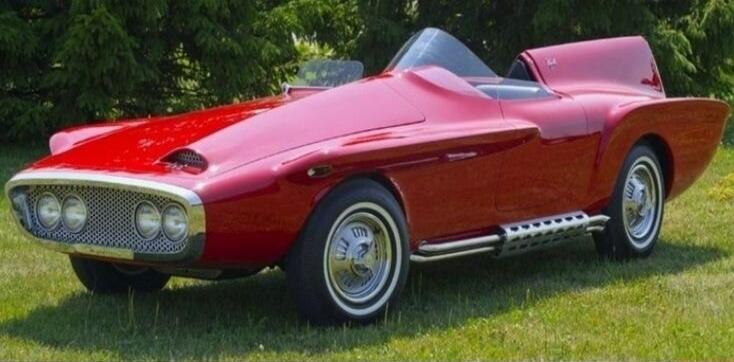 A vintage red concept car with a unique design, featuring multiple headlights and side exhaust pipes, parked on a grassy lawn.