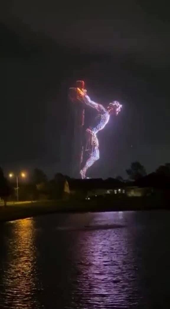 A large, illuminated figure resembling Jesus Christ on a cross is projected in the night sky over a body of water.