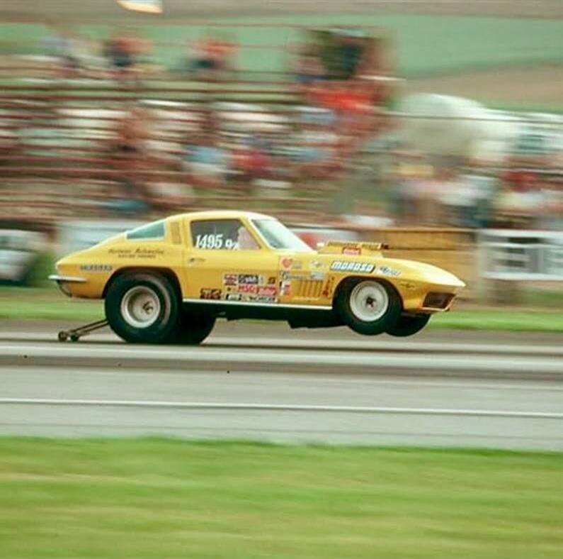 A yellow classic drag racing car, possibly a Corvette C2, performs a wheelie on a drag strip. The car has '1495 9' on its side window and 'MOROSO' on the hood scoop. Spectators are visible in the blurred background.