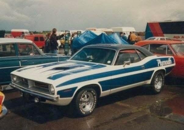 A classic white Plymouth Barracuda muscle car with blue racing stripes is parked outdoors, likely at a car show or event. The word 'Plymouth' is visible on the side of the car. Other vehicles and people are in the background under a cloudy sky.