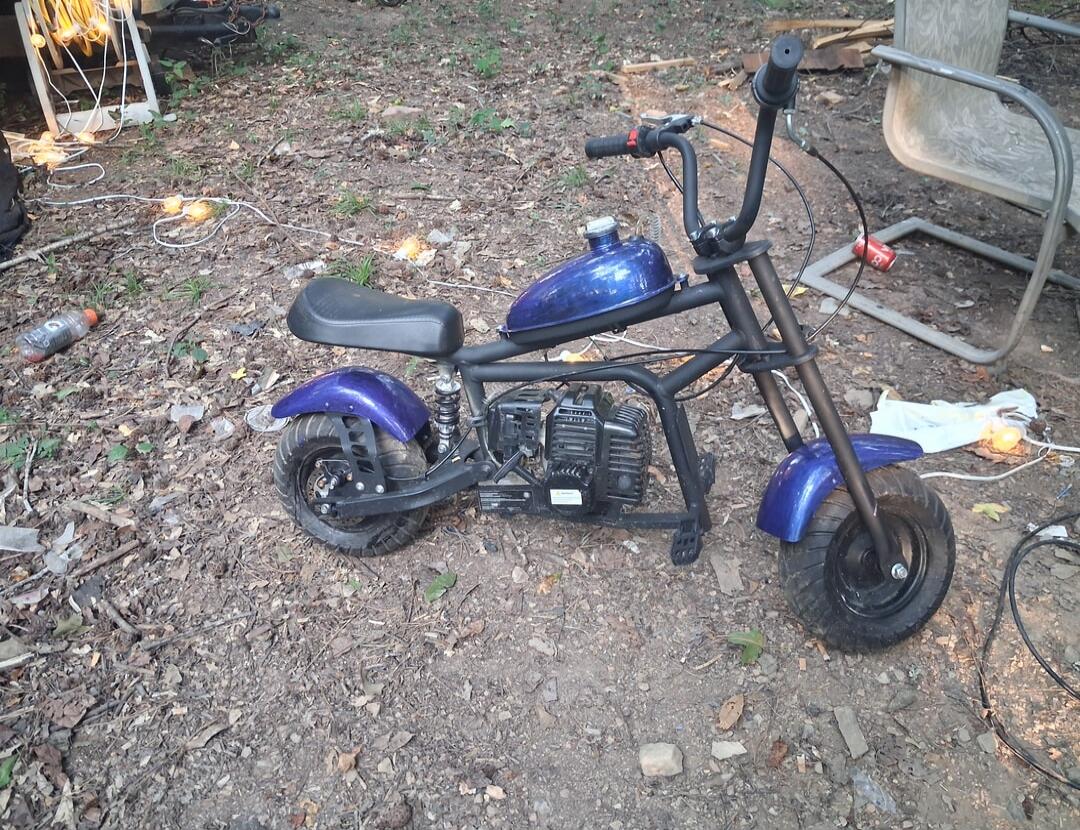 A blue mini bike is parked on a dirt ground outdoors. The bike has a black frame, a blue fuel tank, and blue fenders. There are some leaves, twigs, and string lights scattered on the ground around it.