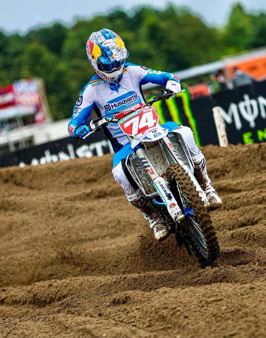 A motocross rider on a Husqvarna bike with number 74, wearing a Bell helmet and Red Bull goggles, racing on a dirt track. Logos for Motorex, Dunlop, and Monster Energy are visible in the background.