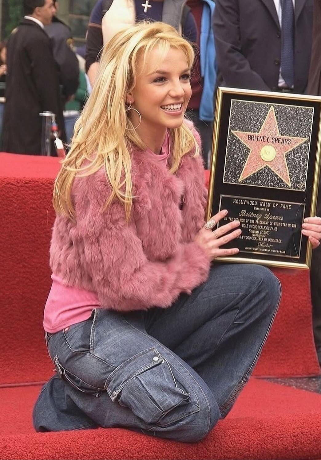BRITNEY SPEARS HOLLYWOOD WALK OF FAME PRESENTED TO BRITNEY SPEARS ON THE OCCASION OF THE PLACEMENT OF YOUR STAR IN THE HOLLYWOOD WALK OF FAME NOVEMBER 17, 2003 HOLLYWOOD CHAMBER OF COMMERCE JOHNNY GRANT CHAIRMAN MAYOR OF HOLLYWOOD. Britney Spears is seen smiling and kneeling on the red carpet, holding her Hollywood Walk of Fame star plaque.