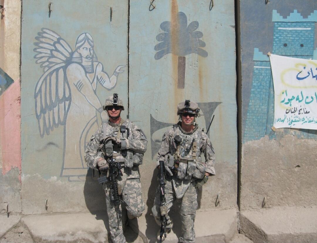 Two soldiers in camouflage uniforms standing in front of a mural on a concrete wall, each holding rifles.
