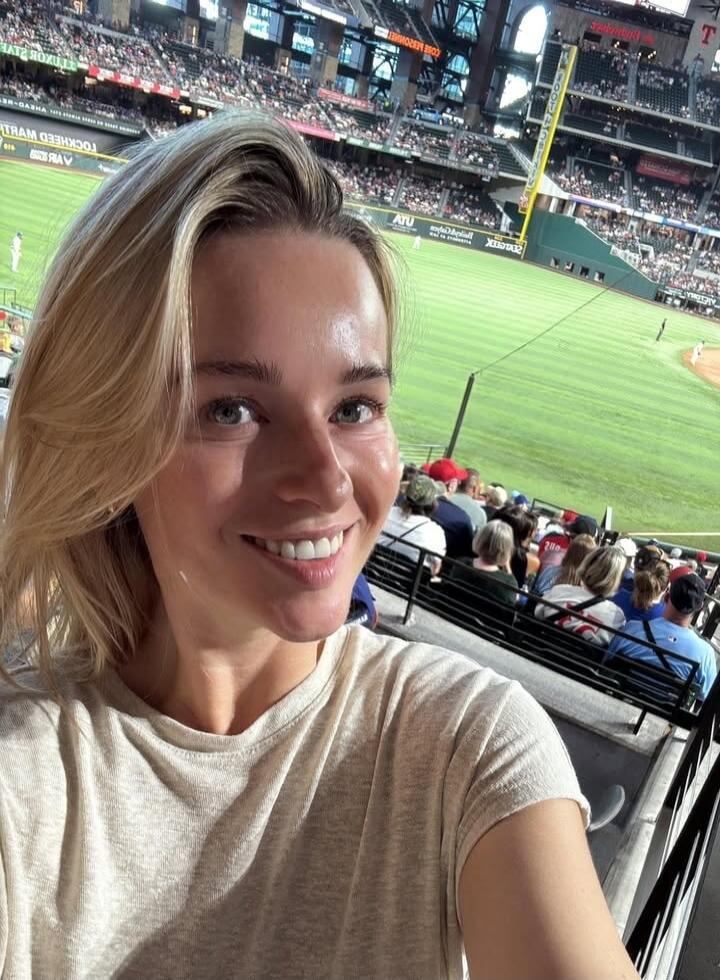 A woman smiling and taking a selfie at a baseball stadium. The stadium features a green field, stands filled with spectators, and various signs, including 'LOCKHEED MARTIN' and 'ATU'.