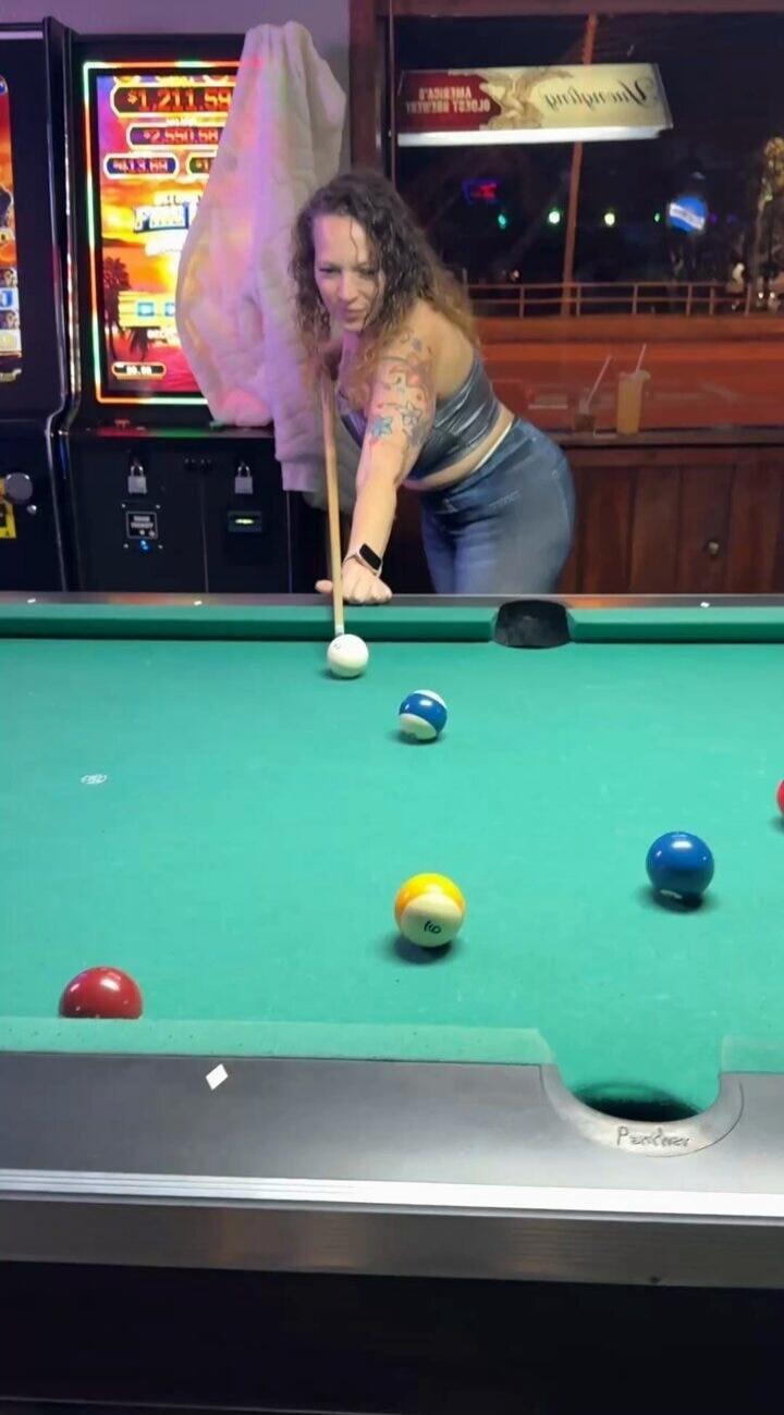 A woman is playing pool in a bar. There are slot machines in the background showing amounts like $1,211.59 and $2,550.58. A sign above the bar reads 'PREMIUM' and 'CANDY'. The pool table has 'Poolover' written on it.