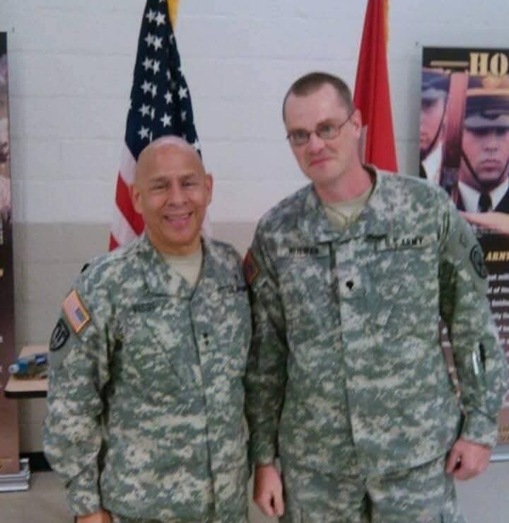 Two men in US Army uniforms are standing. One man's uniform has 'WEST' visible, and the other's has 'WHITMER' and 'U.S. ARMY'. An American flag and a red flag are in the background, along with a poster that partially shows 'HO' and 'ARMY'.
