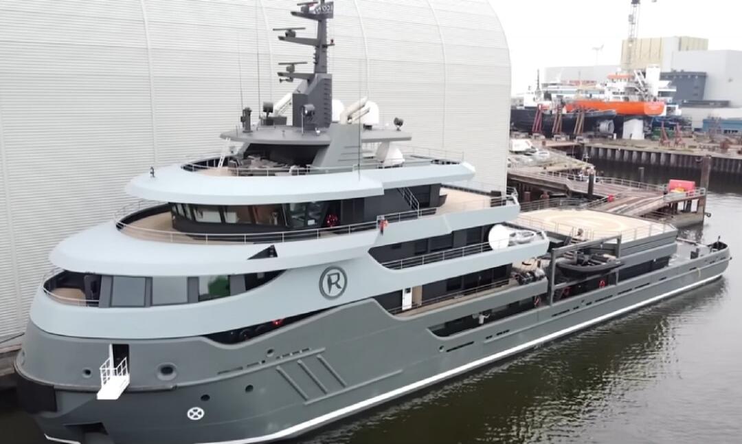 The video shows a large, modern yacht docked at a shipyard. The yacht is grey with white accents and features multiple decks, large windows, and a helipad on the stern. The shipyard appears to be industrial, with cranes and other vessels visible in the background. The overall impression is one of luxury and advanced engineering.