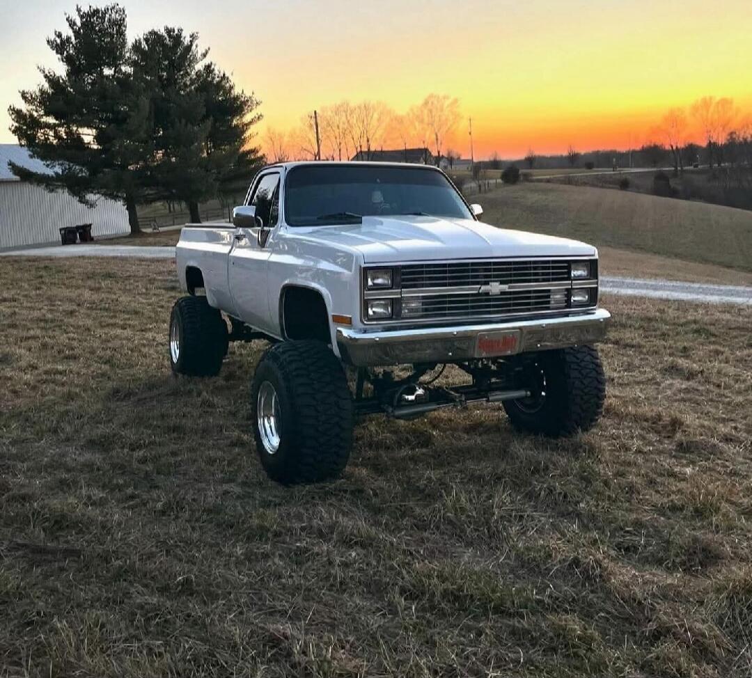 A white lifted pickup truck with large off-road tires parked on a grassy field during sunset.