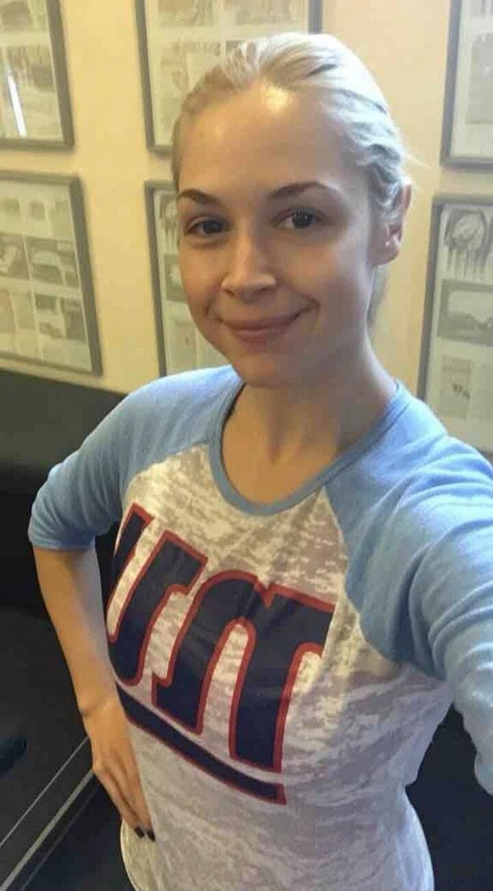 A blonde woman smiling and taking a selfie. She is wearing a light blue and white t-shirt with a red and blue logo that appears to be 'RM' or 'M'. Framed pictures are visible in the background.