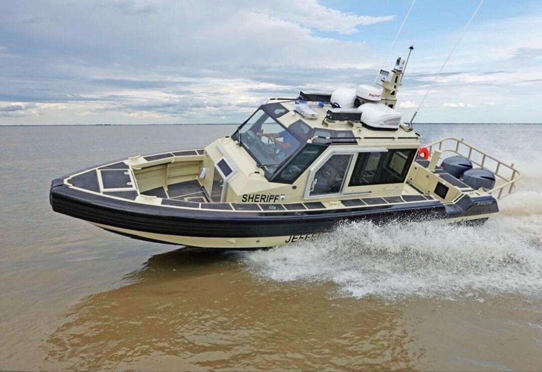 A sheriff's patrol boat, with 'SHERIFF' and 'JEFFER' visible on its side, is moving through murky water under a cloudy sky, creating a wake.