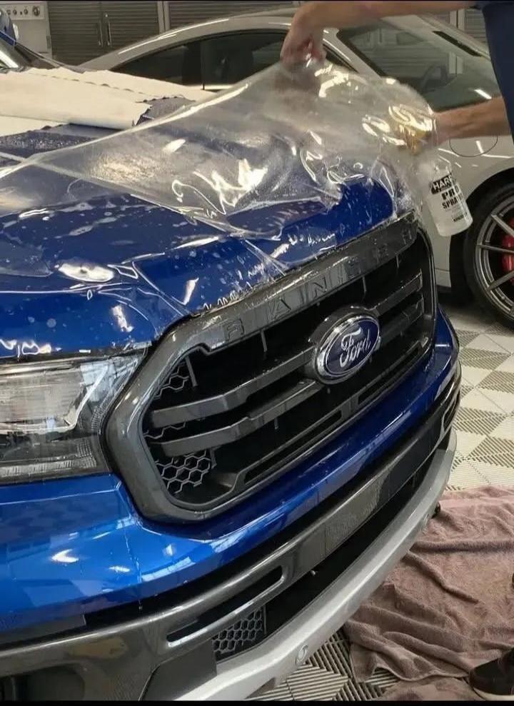 A person is applying a clear protective film to the hood of a blue Ford Ranger pickup truck. The film is being carefully laid over the wet surface, and a spray bottle labeled 'HARD PRO SPRAY' is visible. The process aims to protect the car's paint.