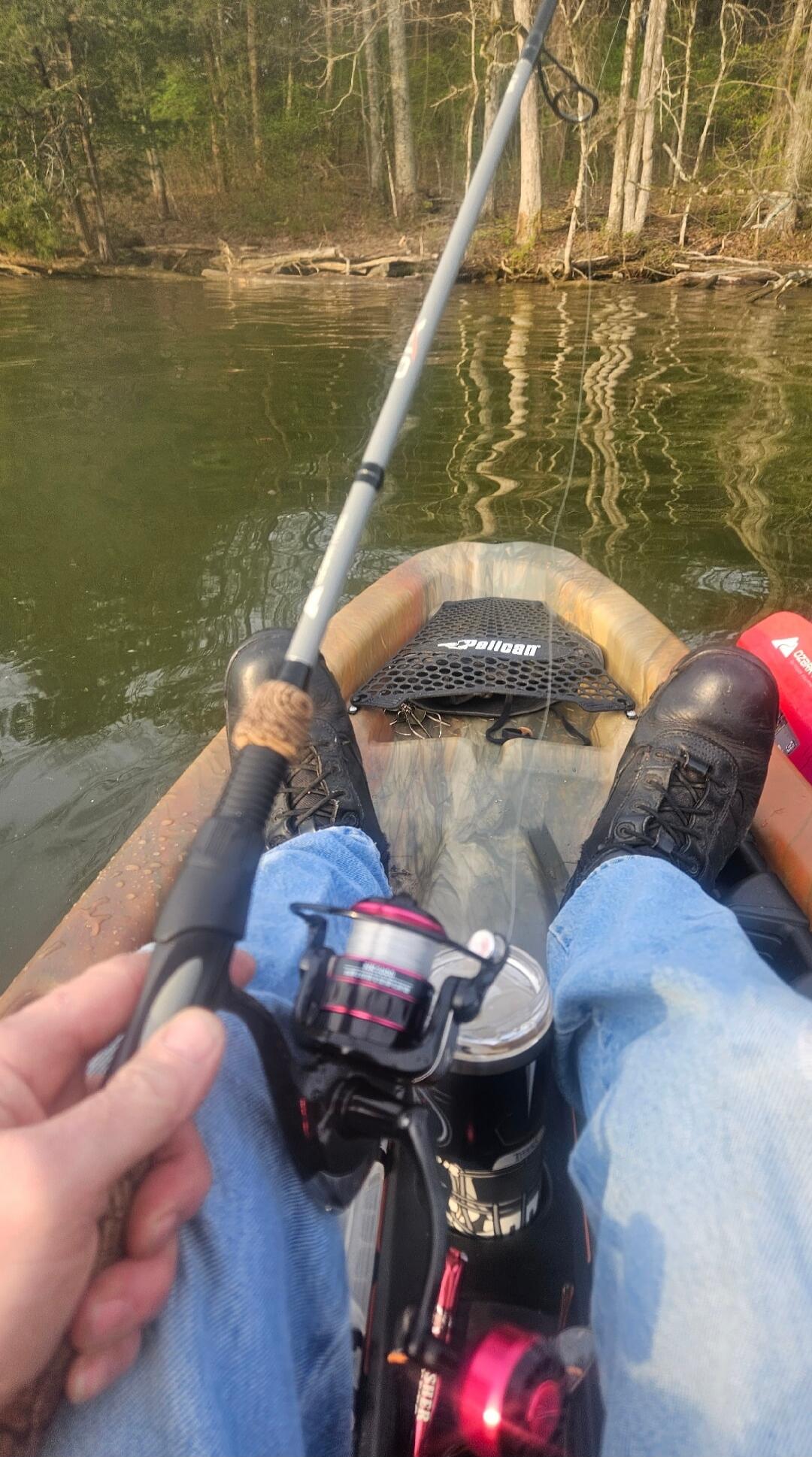 A person is fishing from a kayak on a calm body of water. Trees line the distant shore. The person is holding a fishing rod with a reel, and their legs are visible, wearing jeans and boots. A 'Pelican' brand item is also visible in the kayak.