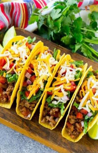 Several hard shell tacos filled with ground beef, shredded cheese, lettuce, diced tomatoes, and red onions, garnished with cilantro and lime wedges.