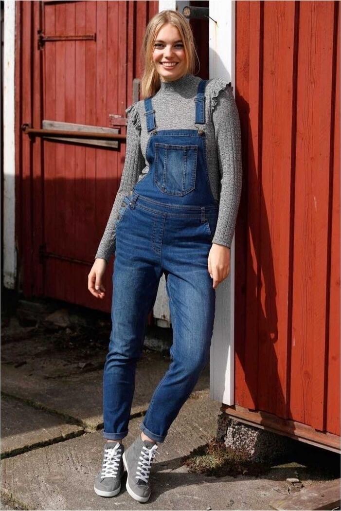 A young woman stands outdoors against a red wooden wall. She is wearing a grey, long-sleeve top under blue denim overalls and grey sneakers with white laces.