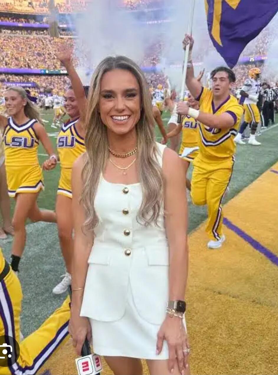A woman in a white sleeveless dress standing on a football field, surrounded by LSU cheerleaders and fans in the stands.