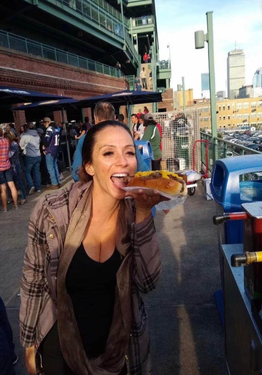A woman with a big smile and her tongue out, holding a large hot dog with mustard and onions, seemingly about to take a bite. She is wearing a plaid jacket over a black top. In the background, there are people, stadium seating, and a city skyline.