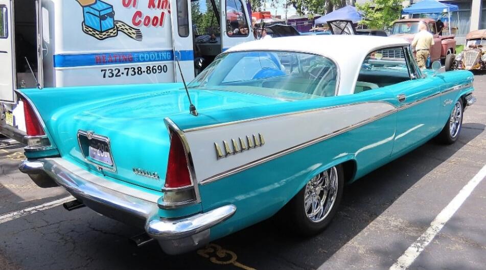 A classic American car, possibly a Chrysler from the late 1950s, with a distinctive teal and white two-tone paint job and chrome details, is parked at an outdoor event. In the background, a white van with 'Kin Cool HEATING & COOLING' and the phone number '732-738-8690' is visible.