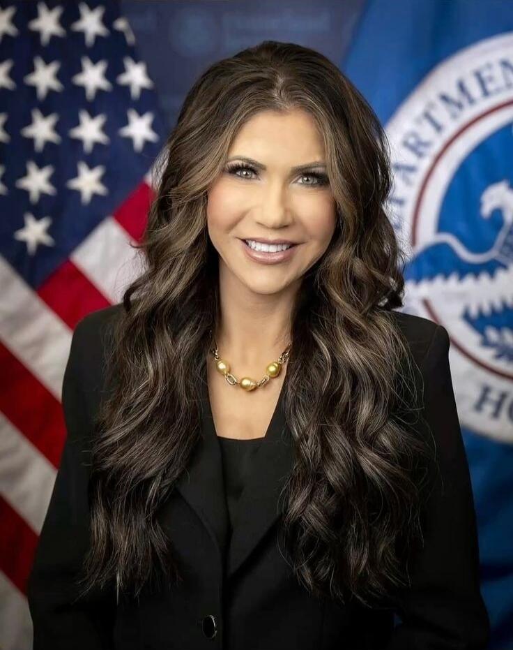 Portrait of a woman in a black blazer with a gold necklace, posed in front of a backdrop featuring the American flag and an official seal.