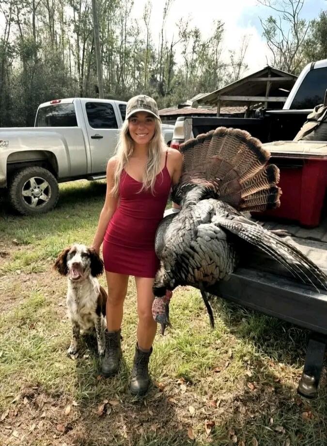 A young woman in a red dress and a cap stands next to a large wild turkey and a dog, with a pickup truck in the background. She is smiling.