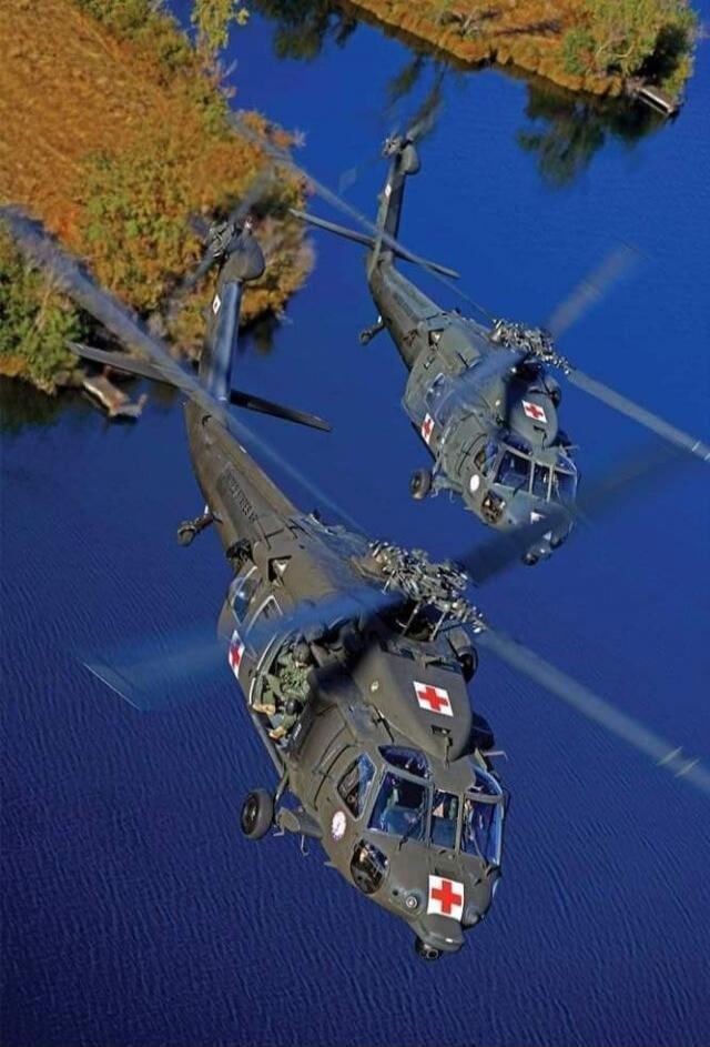 Two military medical helicopters with red cross symbols flying over blue water, with autumn-colored trees along the shore.