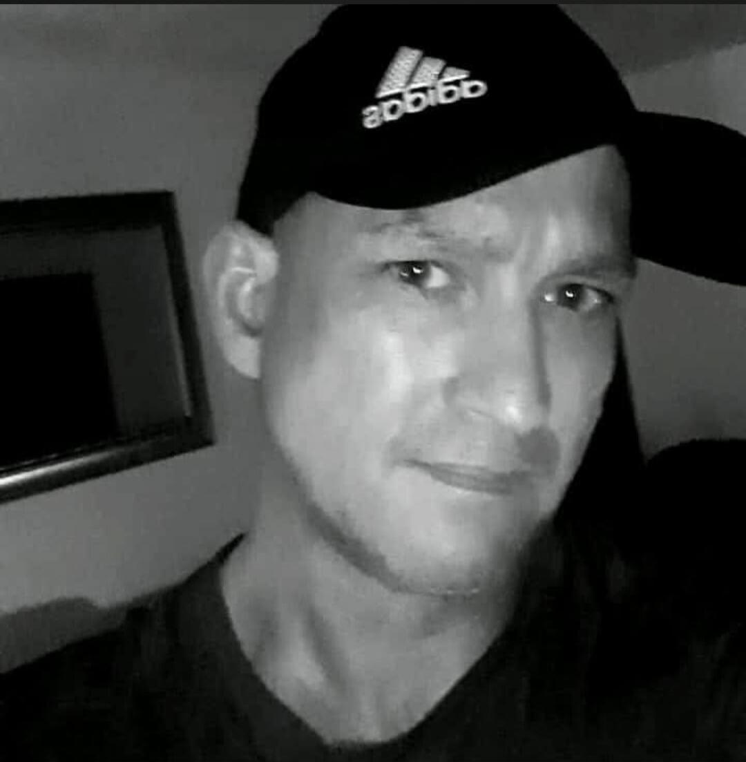 A man wearing a black cap with the Adidas logo (reversed) looks at the camera. The photo is in black and white.