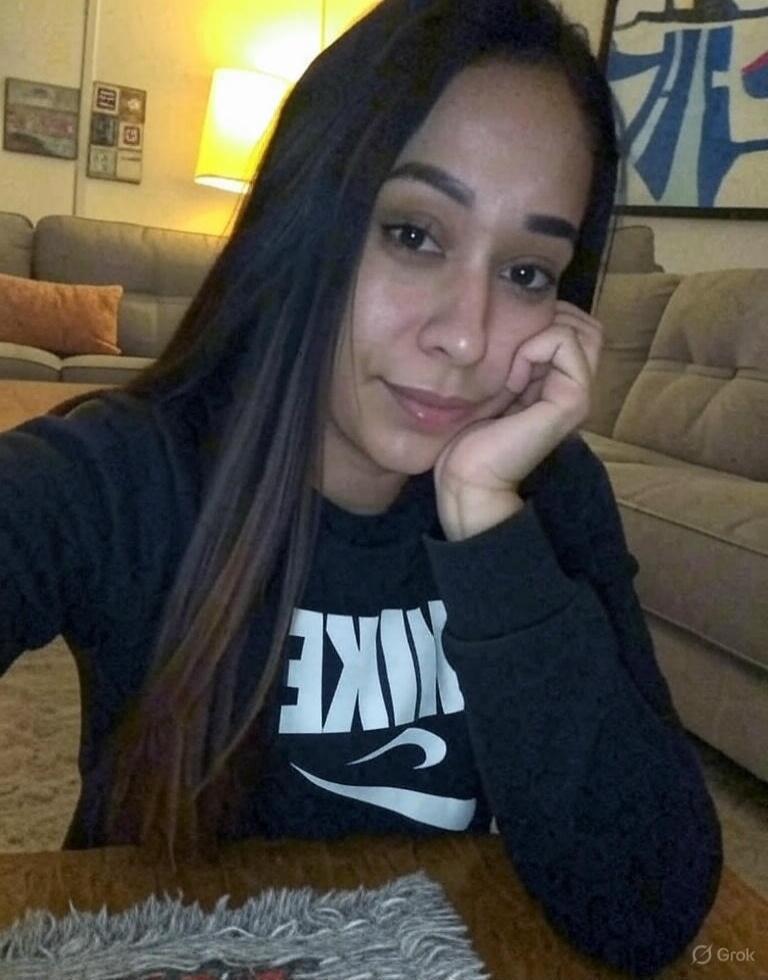 A young woman with long dark hair is looking at the camera, resting her chin on her hand. She is wearing a black sweatshirt with a white 'NIKE' logo (reversed). The background shows a living room with a couch and a lamp.
