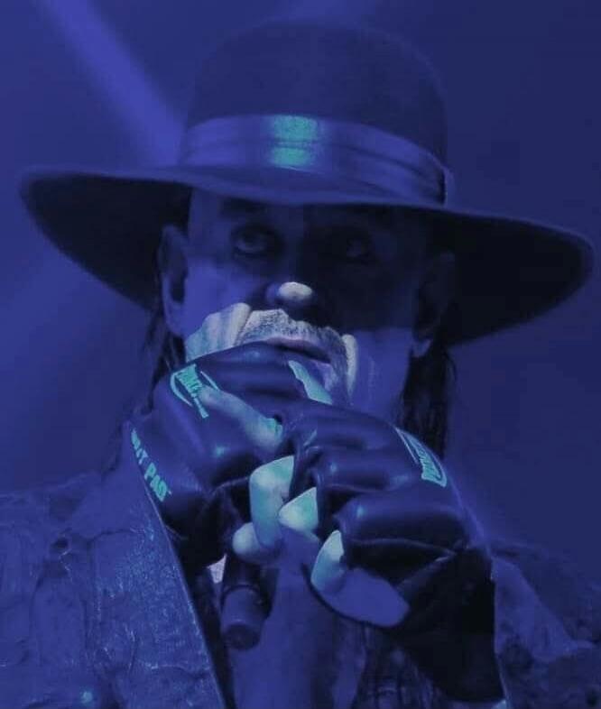 Man wearing a wide-brim hat and gloves, holding a microphone, in blue stage lighting.